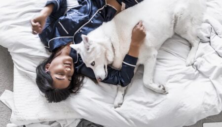 人_白いベッドの上にあおむけになり、白い犬を片手に抱く青いシルクのパジャマを着た黒髪の女性